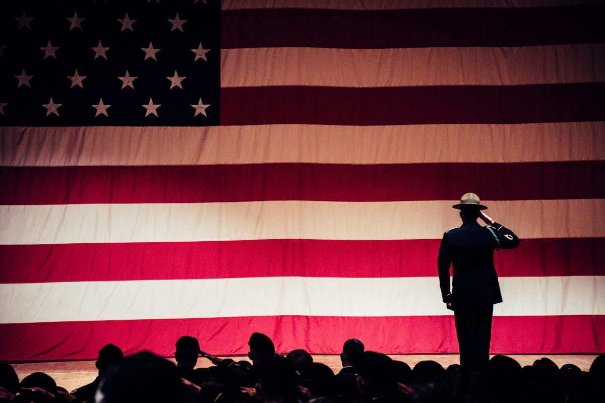 A uniformed service member stands at attention before a large American flag, symbolizing the service that earned the Aid & Attendance benefit