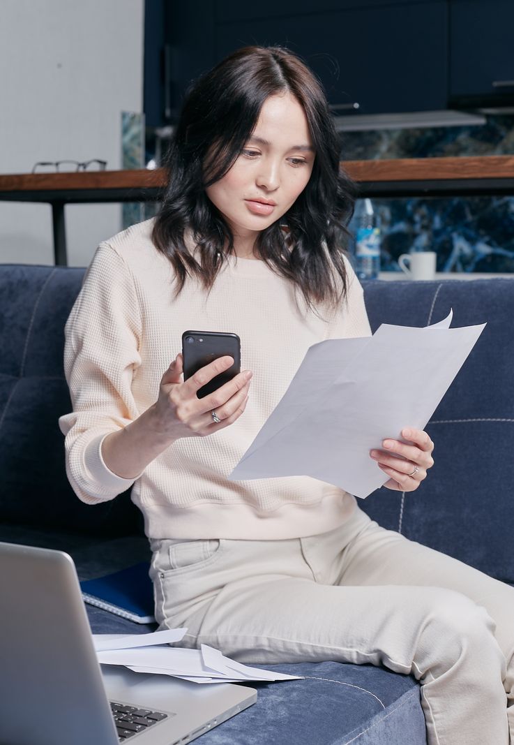 A woman at home reviewing a financial document while holding her phone, with a laptop nearby