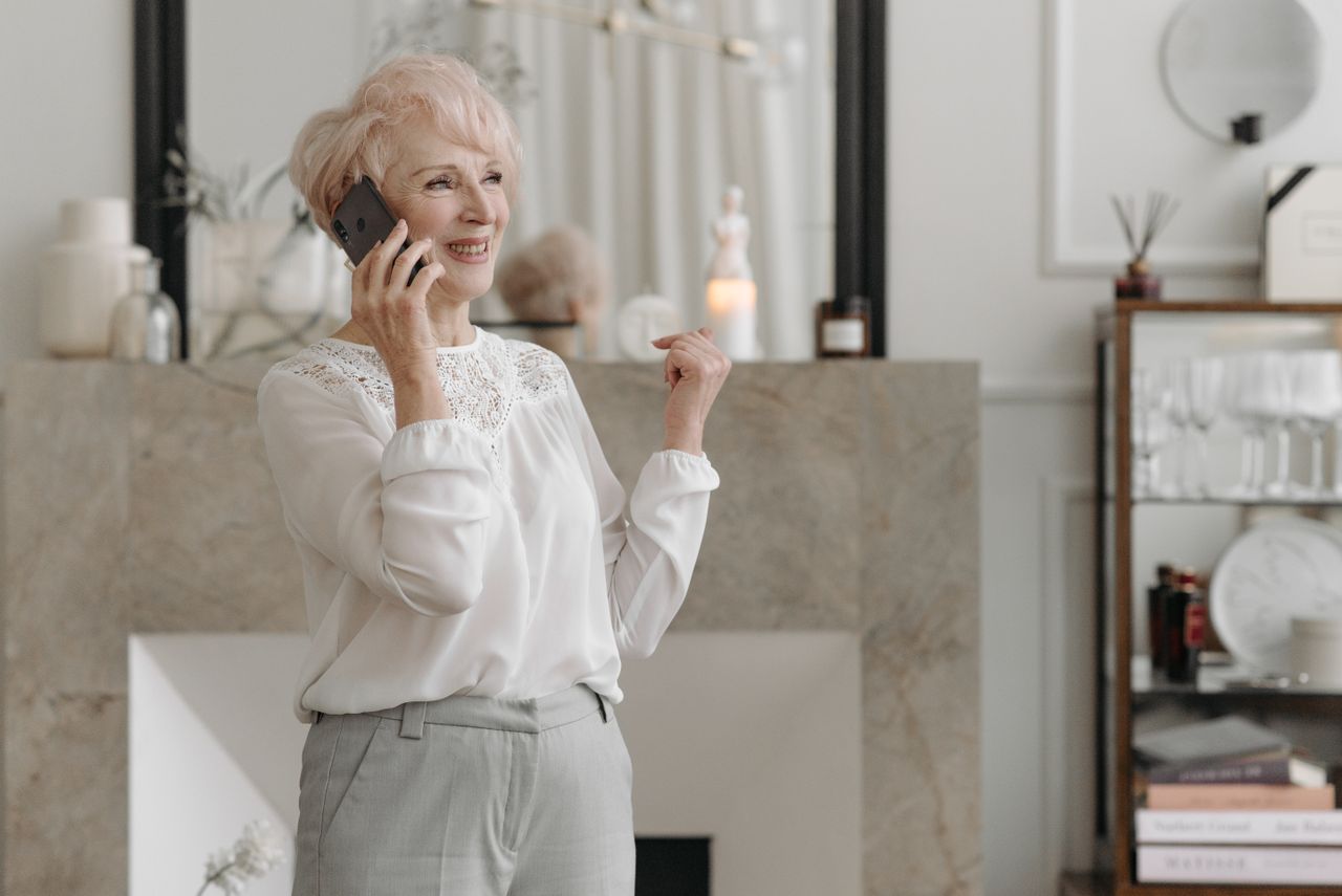 A senior woman smiling while talking on the phone in her living room &mdash; calling to confirm IHSS and Medi-Cal status