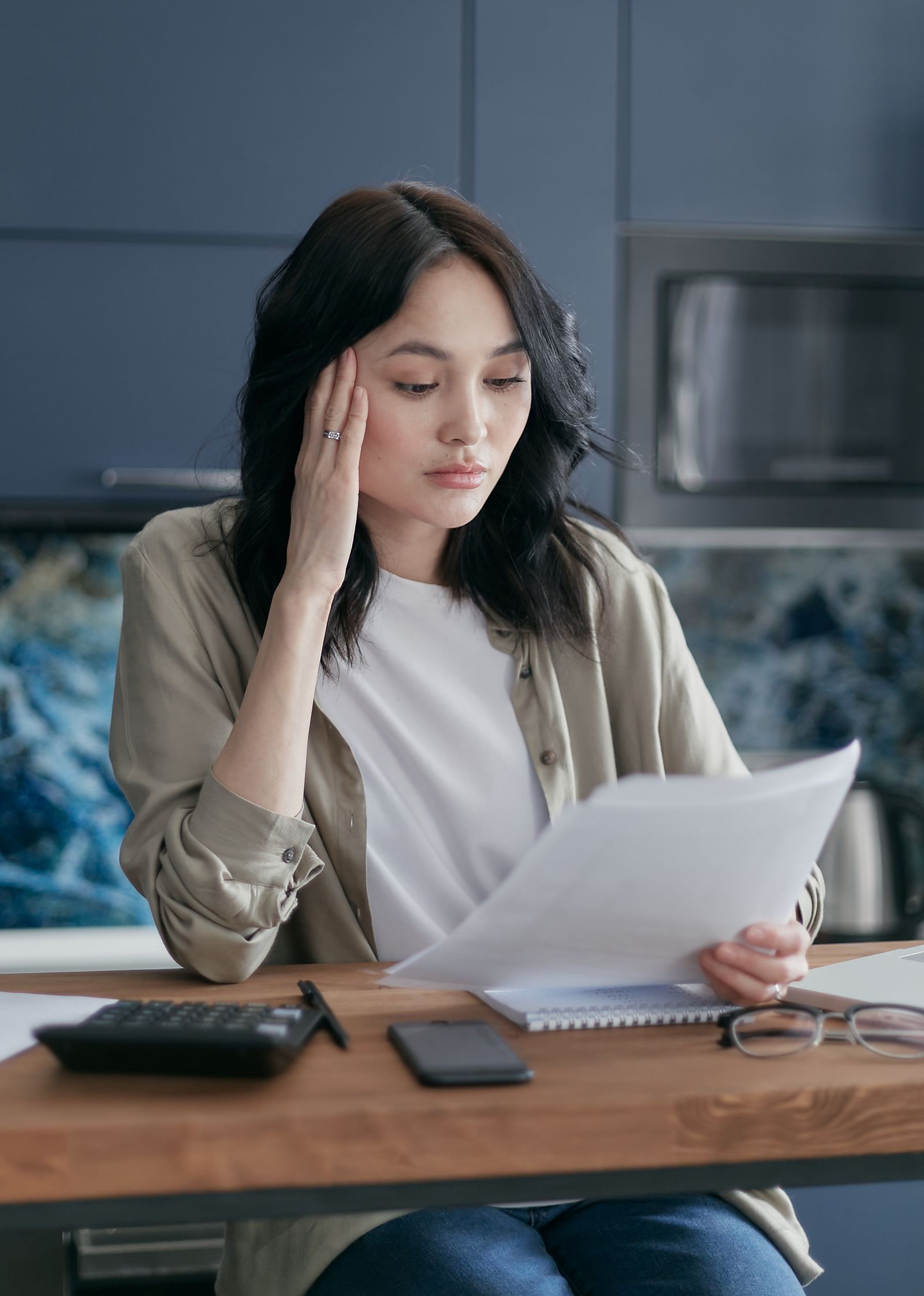 A woman sits at a kitchen table reviewing a printed notice, with a calculator and notepad in front of her