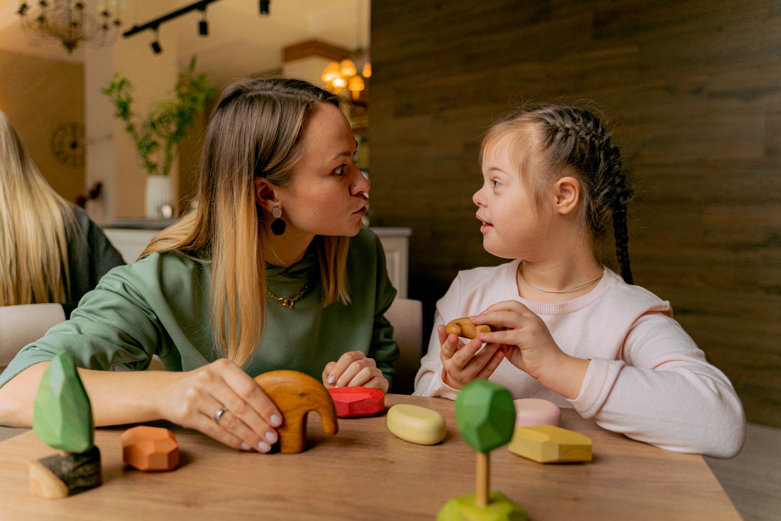 A respite provider sits at a table engaging a young girl with Down syndrome in play with wooden toys
