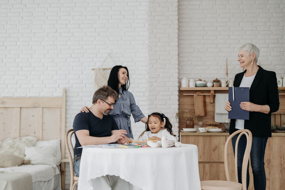 A family sits at a kitchen table while a service coordinator holding a clipboard stands nearby smiling