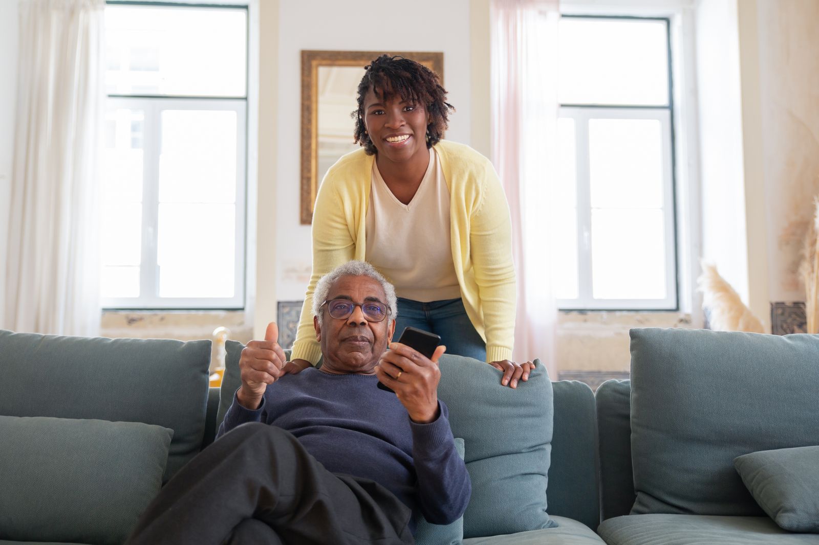 A smiling caregiver stands behind a senior man giving a thumbs up together in a bright living room