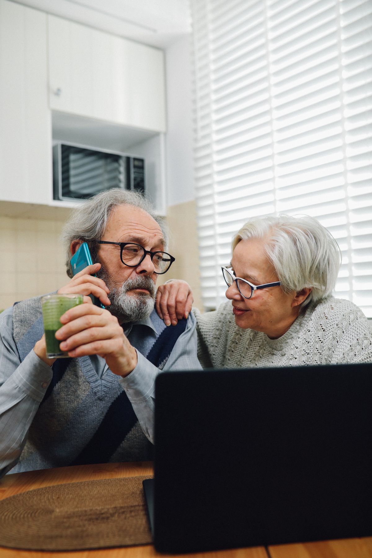 Senior couple making a phone call together while looking at a laptop in their kitchen