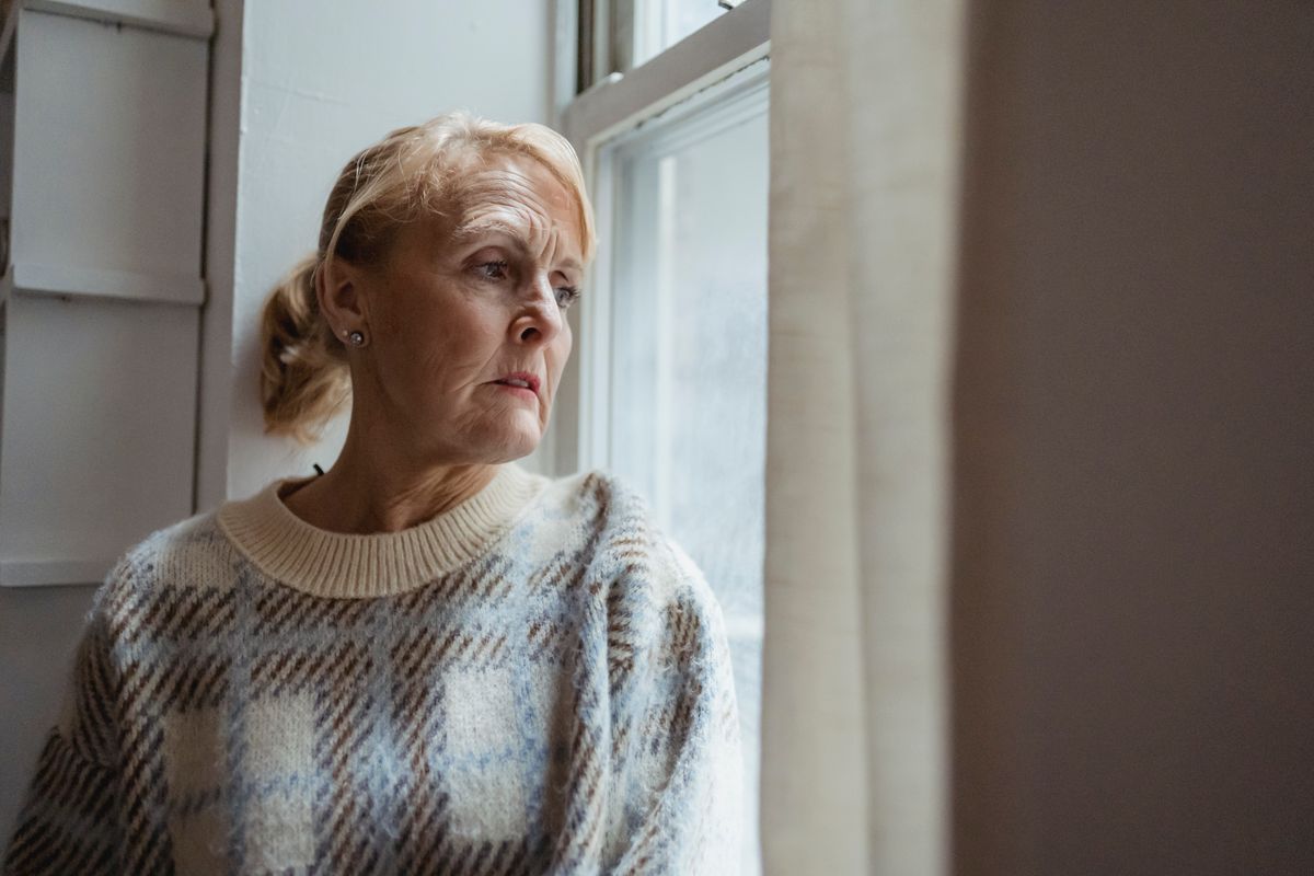 A senior woman looking anxiously out a window in fading evening light, representing sundowning symptoms that worsen as daylight saving time ends