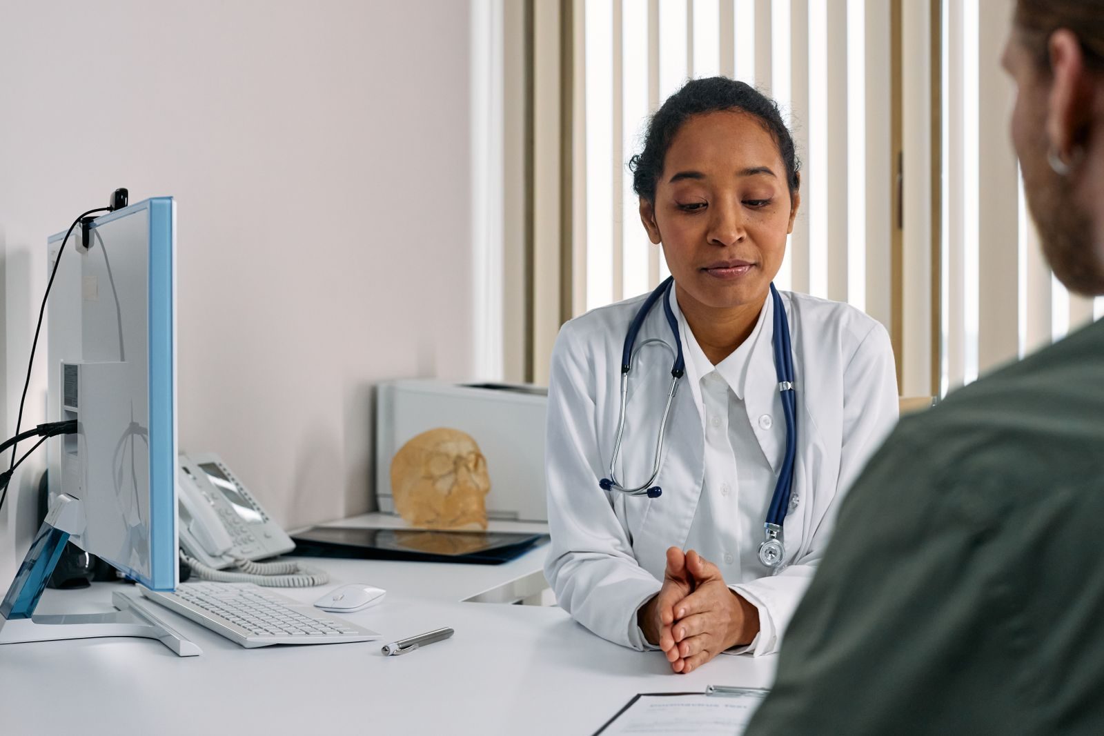 Physician in a white coat with a stethoscope listening to a patient across a desk with a computer monitor