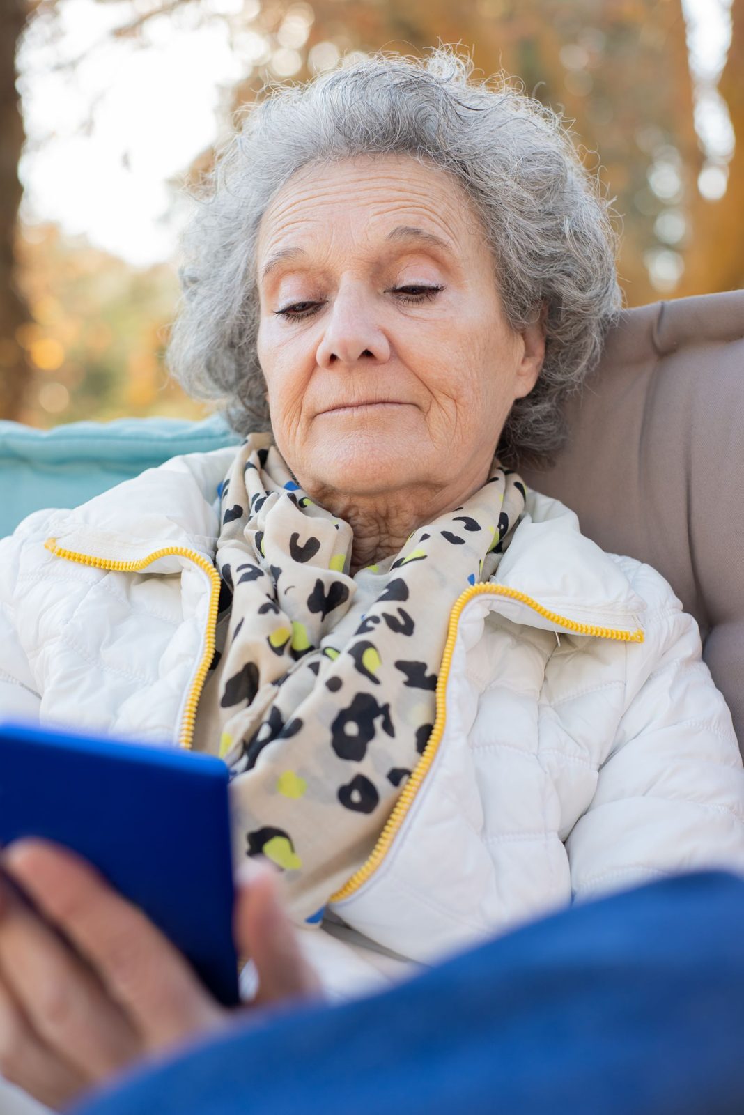 Older woman with gray curly hair in a white jacket and scarf looking at her smartphone
