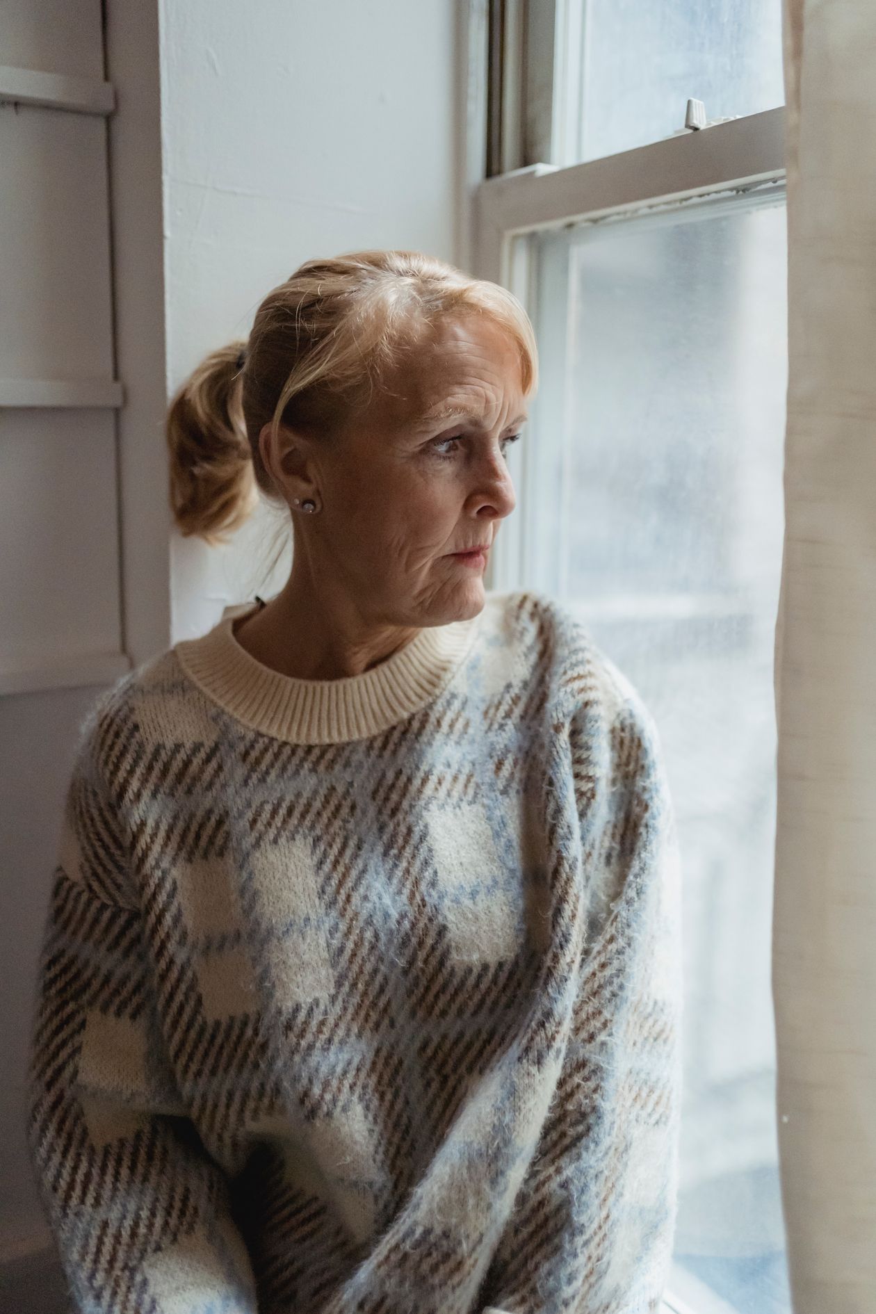Older woman in a sweater looking thoughtfully out a window in soft evening light