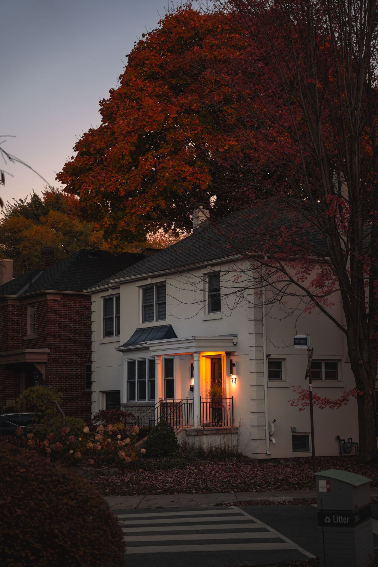 Suburban house with porch light on at dusk during autumn, tree with fall foliage overhead