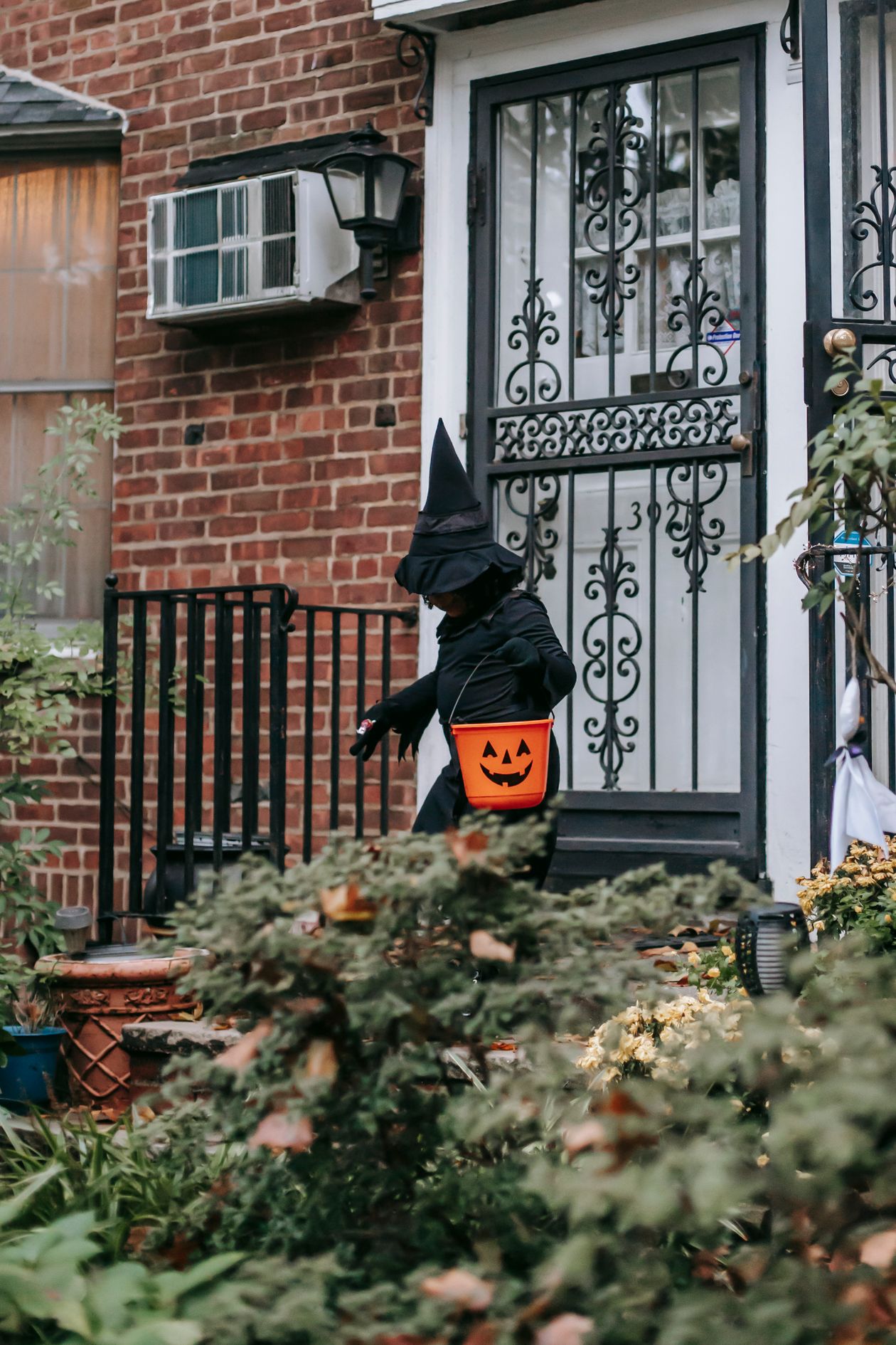 Child in a witch costume carrying a jack-o-lantern candy bucket approaching a front door