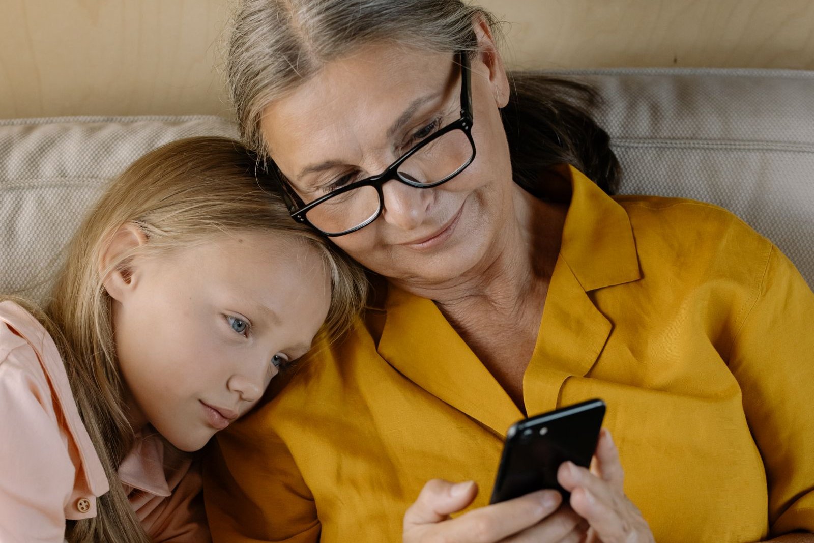 Grandmother in glasses and a mustard-yellow shirt smiling at her phone while her granddaughter rests on her shoulder