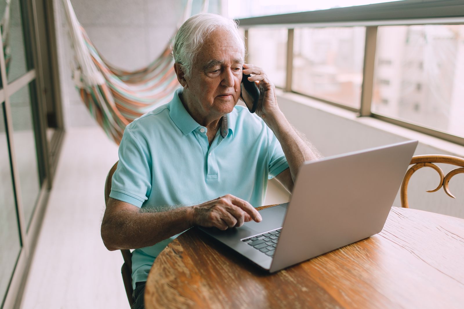 Older man in a light blue polo shirt on a phone call while typing on a laptop at a wooden table