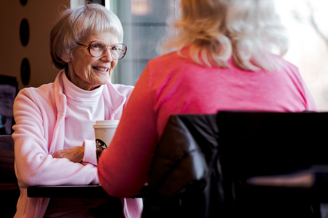 Older woman in a pink sweater smiling across a cafe table at a friend