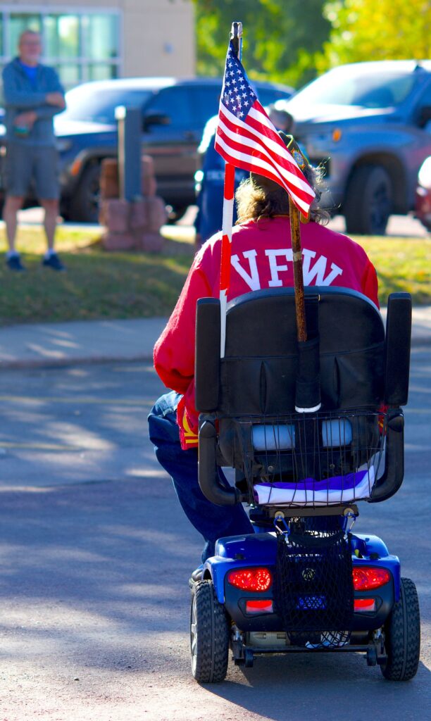 Veteran in a red VFW jacket riding a mobility scooter with an American flag mounted on the back